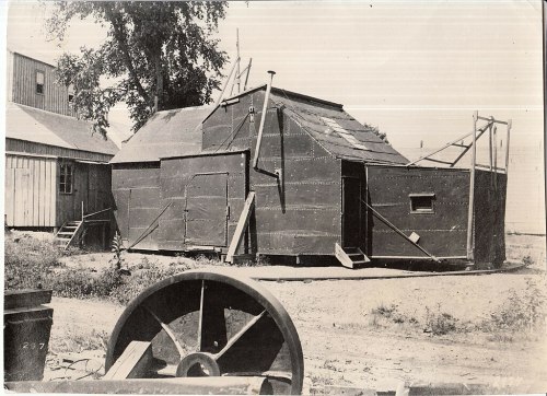 A vintage black-and-white photo of a metal structure with a slanted roof, surrounded by a rural landscape.