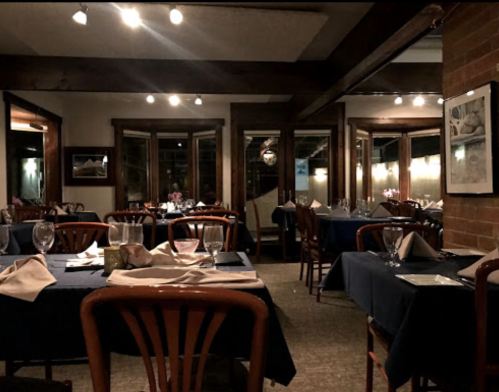Cozy restaurant interior with wooden chairs, blue tablecloths, and dim lighting, featuring large windows and elegant table settings.