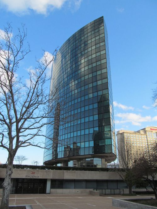 A modern glass skyscraper with a curved design, reflecting clouds and nearby buildings against a blue sky.