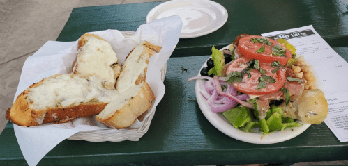 A plate with cheesy garlic bread and a fresh salad topped with tomatoes, olives, and herbs on a green table.