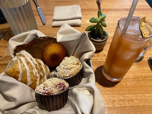 A basket of baked goods including muffins, a pastry, and a cornbread, alongside a drink and a small plant on a wooden table.