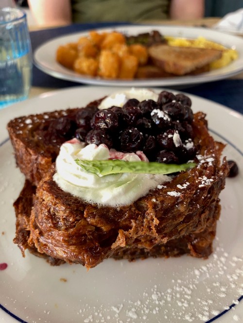 A slice of French toast topped with whipped cream and blueberries, served on a white plate with powdered sugar.