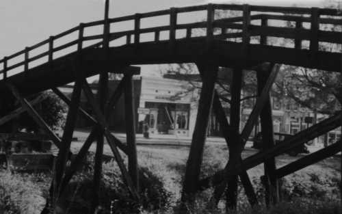 A wooden bridge arches over a grassy area, with a building visible in the background. The image is in black and white.