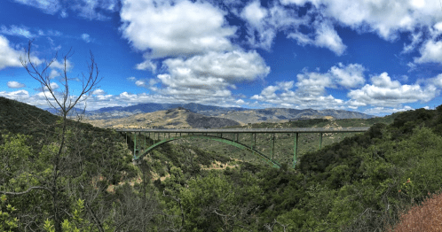 A green bridge spans a valley, surrounded by lush greenery and rolling hills under a partly cloudy sky.