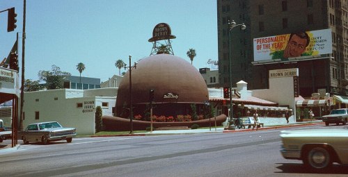 A vintage photo of the Brown Derby restaurant, featuring its iconic hat-shaped building and nearby billboards.