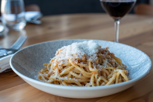 A plate of pasta topped with grated cheese, accompanied by a glass of red wine on a wooden table.
