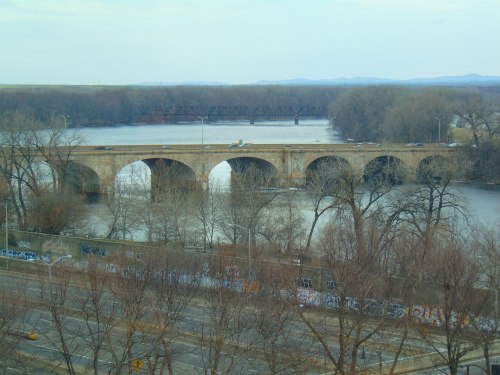 A stone bridge with arches spans a river, surrounded by bare trees and distant hills under a cloudy sky.
