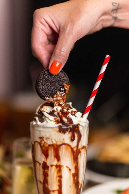 A hand with orange nails dips an Oreo cookie into a tall glass of milkshake topped with whipped cream and chocolate sauce.