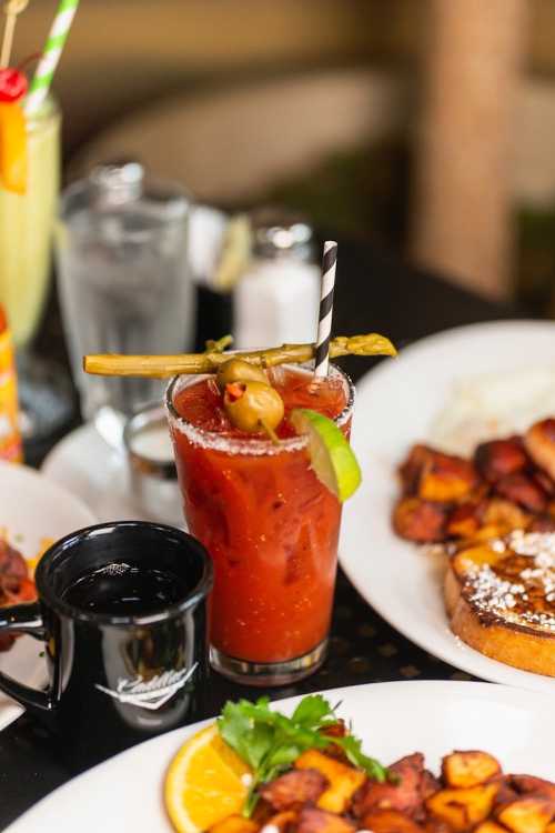 A close-up of a Bloody Mary cocktail garnished with olives and celery, surrounded by breakfast dishes on a table.