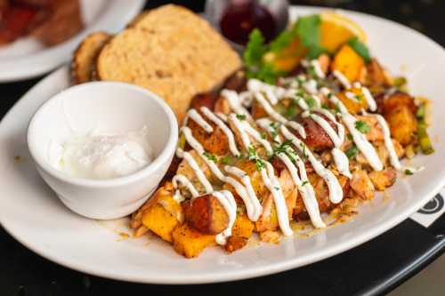 A plate of seasoned potatoes topped with sauce, served with a side of yogurt, toast, and a slice of orange.