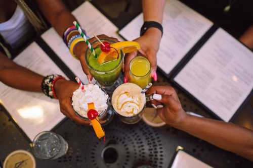 Four colorful cocktails with garnishes are raised in a toast above a table with menus.