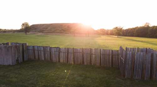 A sunlit landscape featuring a grassy field, a wooden fence, and a distant hill under a clear sky.