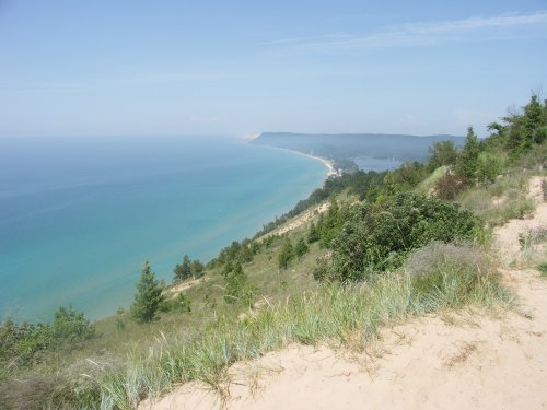 A scenic view of a coastline with sandy dunes, lush greenery, and a calm blue lake under a clear sky.