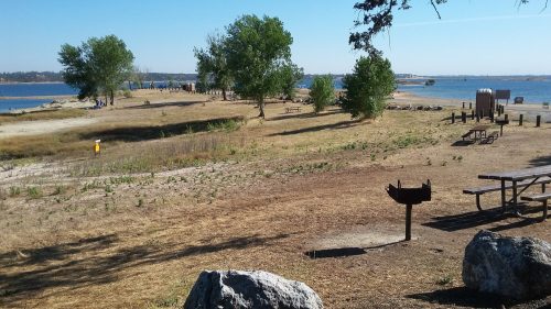 A scenic view of a lakeside park with picnic areas, trees, and a clear blue sky.