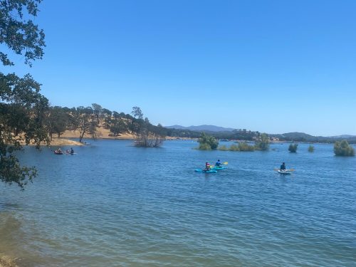 Two kayakers paddle on a calm lake surrounded by trees and hills under a clear blue sky.
