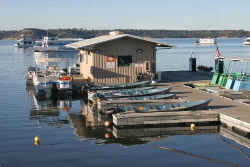 A calm lake scene featuring a boathouse, parked boats, and a clear blue sky reflecting on the water.