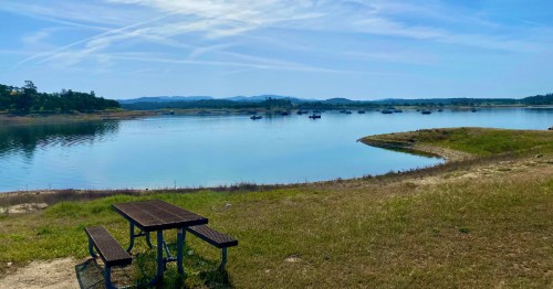 A serene lakeside view with a picnic table, calm water, and distant hills under a clear blue sky.