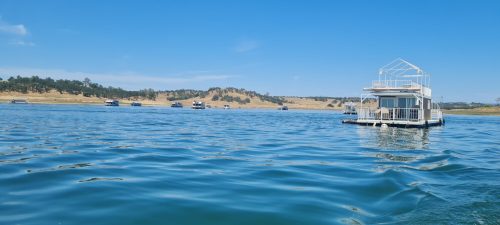 A floating house on calm water, surrounded by boats and a sandy shoreline under a clear blue sky.