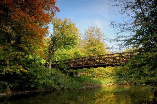 A wooden bridge spans a calm river, surrounded by vibrant autumn foliage and clear blue skies.