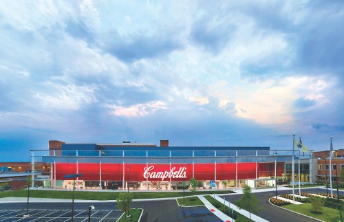 Exterior view of a modern building with a large "Campbell's" sign, surrounded by a parking lot and cloudy sky.