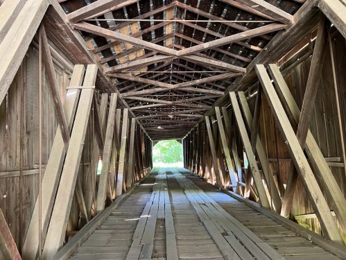 Interior view of a wooden covered bridge, showcasing exposed beams and a wooden floor, with light filtering through.