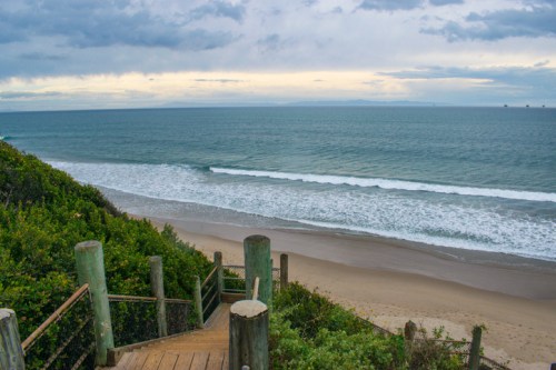 A wooden staircase leads down to a sandy beach with gentle waves and a cloudy sky in the background.