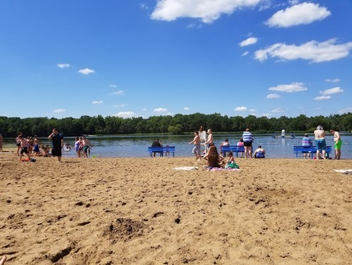 A sandy beach with people enjoying the sun, swimming, and relaxing by a calm lake under a blue sky with clouds.