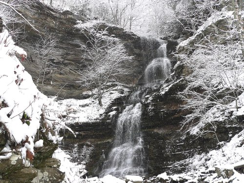 A snowy landscape featuring a cascading waterfall surrounded by rocky cliffs and bare trees.