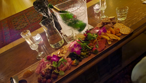 A beautifully arranged charcuterie board with fruits, meats, and flowers, alongside champagne and glasses on a wooden table.