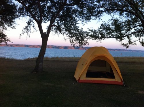 A yellow tent by a lake, surrounded by trees, with a scenic view of hills in the background at dusk.