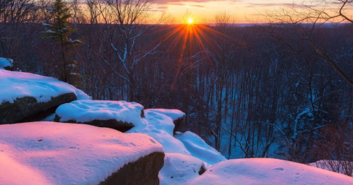 A winter sunset over a snowy forest, with vibrant colors illuminating the sky and glistening on the snow-covered rocks.