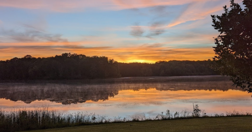 A serene lake at sunrise, reflecting colorful skies and trees along the shore, with mist rising from the water.