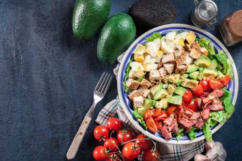 A colorful salad with chicken, avocado, tomatoes, and bacon, surrounded by fresh ingredients on a dark background.