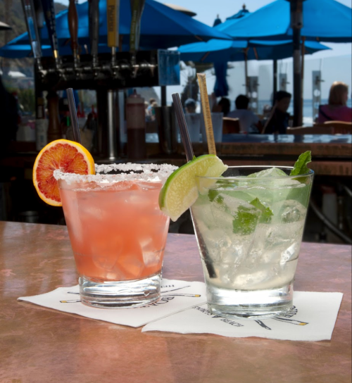 Two colorful cocktails on a copper table: a pink drink with a blood orange slice and a clear drink with mint and lime.
