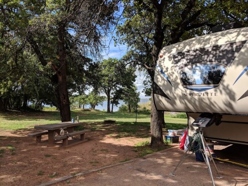 A camper parked at a campsite with a picnic table and trees, overlooking a serene lake in the background.