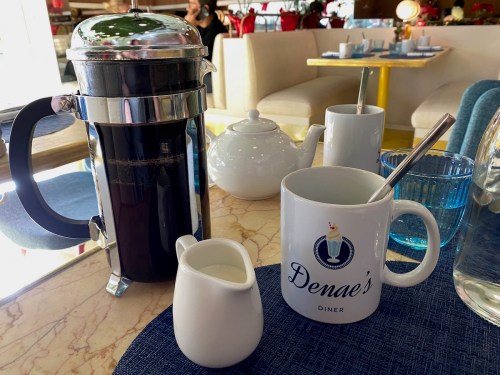 A coffee pot, cream pitcher, and a mug on a table in a diner setting, with soft seating in the background.