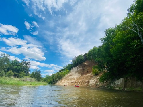 A serene river scene with a sandy cliff and lush greenery under a bright blue sky with fluffy clouds.