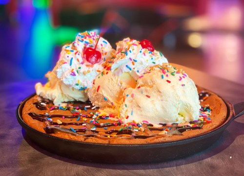 A warm cookie skillet topped with scoops of vanilla ice cream, whipped cream, sprinkles, and cherries.