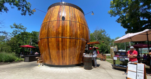 A large wooden barrel structure serves as a kiosk in a sunny outdoor setting with tables and umbrellas nearby.