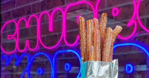 A close-up of churros in a silver wrapper, with a colorful neon sign in the background.