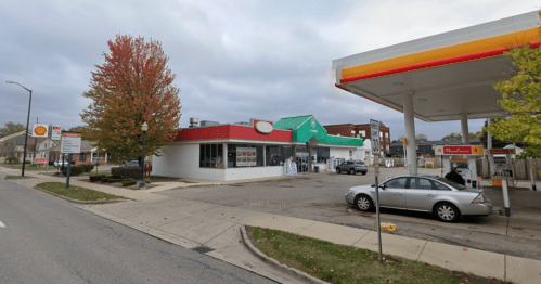 A street view of a gas station and a restaurant with a green roof, surrounded by trees and cloudy skies.