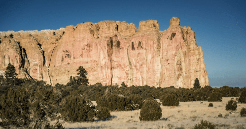 A rugged rock formation rises above a desert landscape, surrounded by sparse vegetation under a clear blue sky.