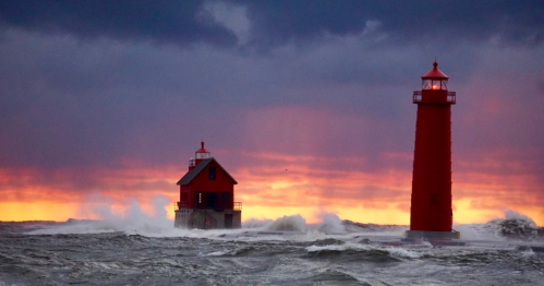 A red lighthouse and a nearby building stand against a stormy sea, with dramatic clouds and a colorful sunset in the background.
