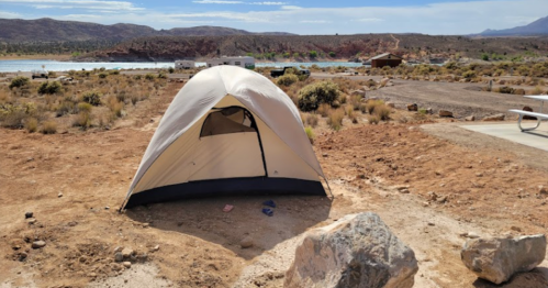 A tent set up on rocky ground near a lake, surrounded by sparse vegetation and mountains in the background.