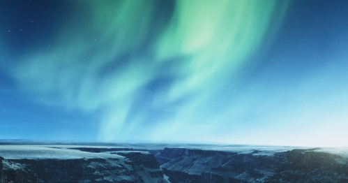 A stunning view of the northern lights illuminating a snowy landscape and canyon under a starry sky.