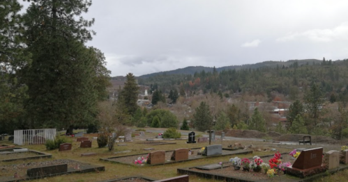 A serene cemetery with gravestones and flowers, surrounded by trees and hills under a cloudy sky.