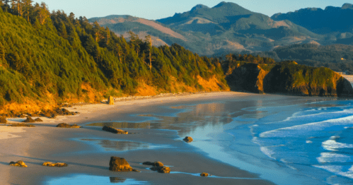 Scenic beach view with rocky shoreline, lush green hills, and calm waves under a clear blue sky.