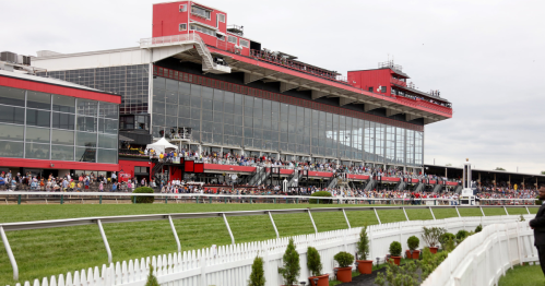 Crowd gathered at a racetrack with a large grandstand and red building in the background, surrounded by greenery.