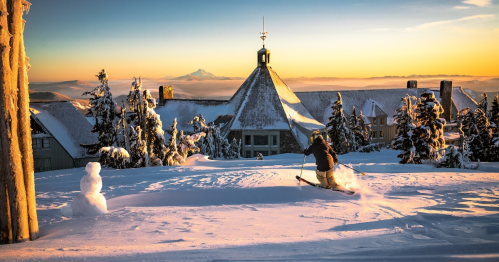 A skier glides through fresh snow at sunset, with a cozy lodge and snowman in the background.