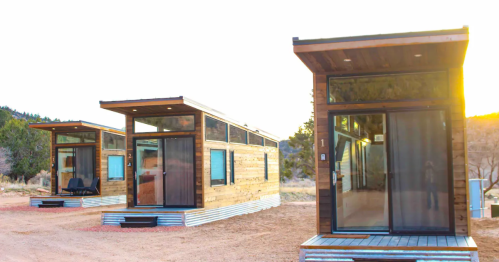 Three modern tiny houses with wooden exteriors and large glass doors, set in a natural landscape at sunset.